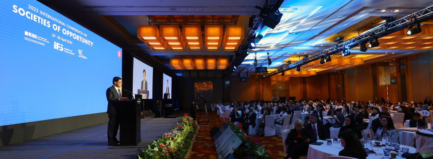 Conference hall with speaker at lectern, large screen displaying 'Societies of Opportunity' and audience seated at tables.